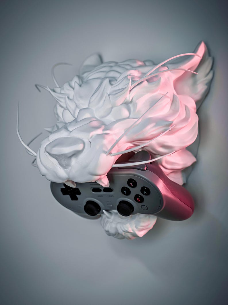 K&ouml;nnte beinhalten: Eine wei&szlig;e Skulptur eines Drachenkopfes, der einen grau-rosa Videospiel-Controller h&auml;lt. Die Skulptur hat detaillierte Texturen und wird mit rosa und wei&szlig;em Licht beleuchtet.