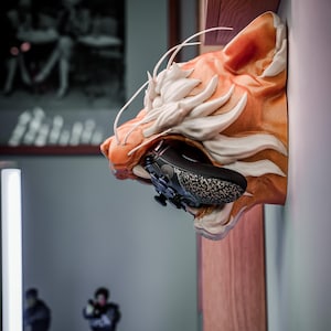 K&ouml;nnte beinhalten: Eine Wandhalterung in Form eines orange-wei&szlig; gestreiften Tigerkopfes, der einen schwarzen, gemusterten Videospiel-Controller h&auml;lt. Die Skulptur ist auf einem Holzregal montiert, mit unscharfem Hintergrund.