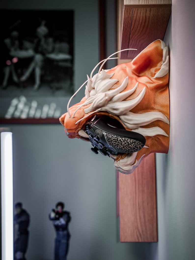 K&ouml;nnte beinhalten: Eine Wandhalterung in Form eines orange-wei&szlig; gestreiften Tigerkopfes, der einen schwarzen, gemusterten Videospiel-Controller h&auml;lt. Die Skulptur ist auf einem Holzregal montiert, mit unscharfem Hintergrund.