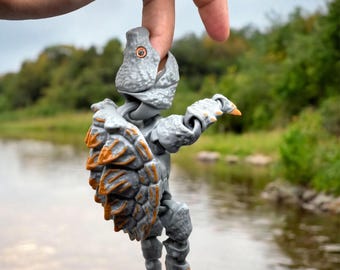 May include: A gray and orange articulated action figure of a creature, held by a hand. The figure has a textured, armored appearance with orange accents on its back and claws. The background shows a river and trees.
