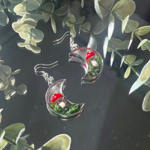 May include: Silver-toned crescent moon earrings. Each earring features a miniature landscape with green moss and a red mushroom with white spots. The earrings are suspended from a simple hook.