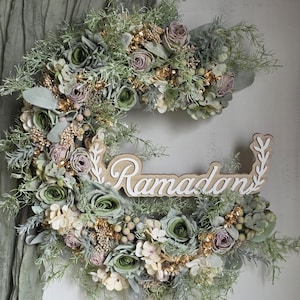 May include: A crescent-shaped floral wreath with the word "Ramadan" in white script. The wreath features green, cream, and mauve flowers, gold accents, and greenery. The background is a light-coloured wall with a green fabric.