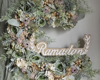 May include: A crescent-shaped floral wreath with the word "Ramadan" in white script. The wreath features green, cream, and mauve flowers, gold accents, and greenery. The background is a light-coloured wall with a green fabric.