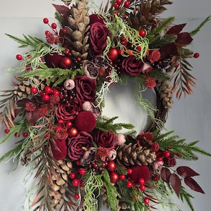 Op de afbeelding: Een decoratieve krans met bordeauxrode rozen, rode bessen en dennenappels. Groen en bruin blad zijn in het ontwerp verwerkt, met rode ballen voor een feestelijke touch. Een seizoensgebonden decoratie.