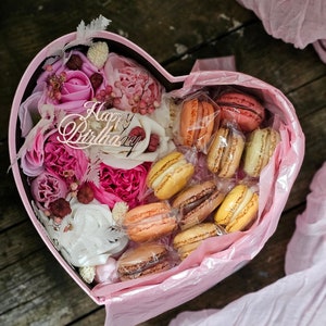 May include: A pink heart-shaped box filled with pink and white flowers and macarons. The box has a sign that says "Happy Birthday".