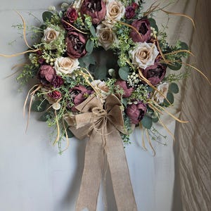 Könnte beinhalten: Ein floraler Kranz mit burgunderfarbenen Pfingstrosen, cremefarbenen Rosen und grünem Laub. Der Kranz ist mit Zweigen goldfarbener Stiele und einer großen Schleife aus Jute verziert. Dieser dekorative Kranz ist für den Innen- und Außenbereich geeignet.