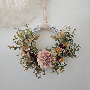 May include: A white wicker wreath decorated with pink and burgundy flowers and green eucalyptus leaves.