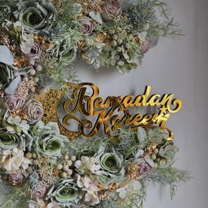 May include: A floral wreath with a gold-coloured "Ramadan Kareem" sign. The wreath features green, white, and mauve-coloured flowers and foliage. The sign is in a cursive font and is placed in the centre of the wreath.
