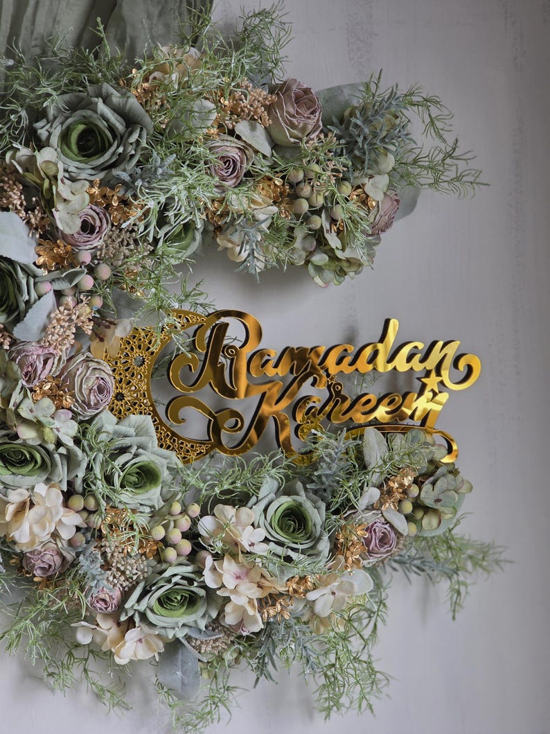 May include: A floral wreath with a gold-coloured "Ramadan Kareem" sign. The wreath features green, white, and mauve-coloured flowers and foliage. The sign is in a cursive font and is placed in the centre of the wreath.