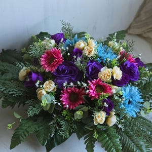 Könnte beinhalten: Ein lebendiger Blumenstrauß mit einer Mischung aus bunten Blumen. Der Strauß enthält lila Rosen, rosa Gerbera, blaue Astern und cremefarbene Blüten, akzentuiert mit grünem Laub. Die Anordnung befindet sich vor einem neutralen Hintergrund.