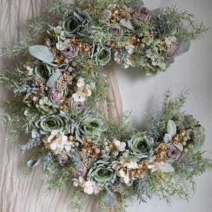 May include: A crescent-shaped floral wreath with green roses, lavender buds, and white flowers. The wreath is decorated with gold-coloured sprigs and various green foliage. The arrangement hangs against a neutral-toned fabric backdrop.