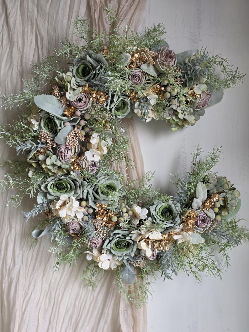 May include: A crescent-shaped floral wreath with green roses, lavender buds, and white flowers. The wreath is decorated with gold-coloured sprigs and various green foliage. The arrangement hangs against a neutral-toned fabric backdrop.