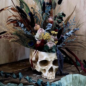 May include: A white skull planter filled with dried flowers and feathers. The flowers are in shades of pink, yellow, and blue. The feathers are brown, black, and green. The skull is sitting on a wooden surface.