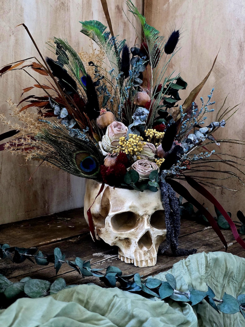 May include: A white skull planter filled with dried flowers and feathers. The flowers are in shades of pink, yellow, and blue. The feathers are brown, black, and green. The skull is sitting on a wooden surface.