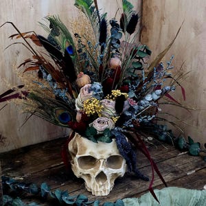 May include: A white human skull with a bouquet of dried flowers in the eye sockets. The flowers include dried roses, eucalyptus, and peacock feathers. The skull is sitting on a wooden surface.