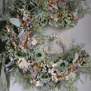 May include: A floral wreath with a wooden "Eid Mubarak" sign. The wreath features green, mauve, and cream-coloured flowers, with gold accents and greenery. The arrangement is set against a neutral background.