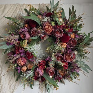 May include: A circular wreath featuring burgundy, red, and peach roses, complemented by green foliage, sprigs, and gold baubles. The wreath's varied textures and colours create a festive, decorative display, suitable for seasonal home decor.