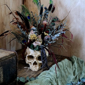 May include: A white skull vase filled with a bouquet of dried flowers, including blue, green, and brown feathers, and dried roses. The vase is sitting on a wooden surface with a green fabric in the background.