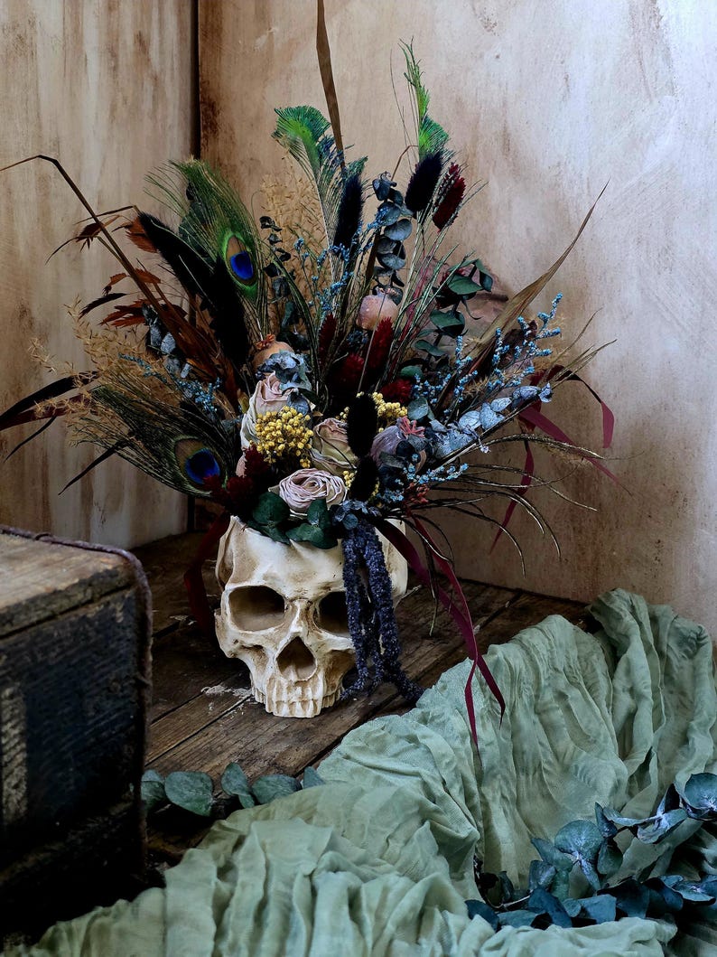 May include: A white skull vase filled with a bouquet of dried flowers, including blue, green, and brown feathers, and dried roses. The vase is sitting on a wooden surface with a green fabric in the background.