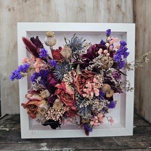 Könnte beinhalten: Eine quadratische Schattenbox mit weißem Rahmen, die ein Trockenblumenarrangement enthält. Die Blumen sind ein Mix aus Lila, Rosa und Rot mit getrockneten Samenkapseln und Gräsern.