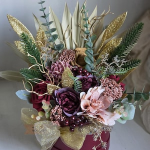 May include: A burgundy round box overflowing with a floral arrangement. The arrangement features burgundy, pink, and cream-colored flowers, accented with gold glitter leaves and a gold ribbon. Greenery and eucalyptus leaves add texture.