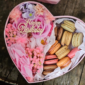 May include: A pink heart-shaped box filled with pink and white flowers and macarons. The box has a gold inscription that reads "Happy Birthday".