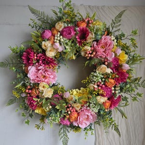 May include: A vibrant floral wreath featuring a variety of colorful blooms. The wreath includes pink peonies, yellow and orange roses, and green foliage. The arrangement is full and lush, perfect for adding a touch of spring to any space.