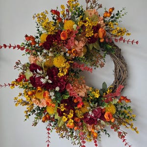 Peut inclure: Une couronne circulaire composée d'une base en brindilles tressées brunes, ornée d'un arrangement de fleurs et de feuillages artificiels dans des tons de bordeaux profond, jaune vif, orange brûlé et pêche, avec des baies et des feuilles vertes.