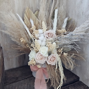 Op de afbeelding: Een boeket gedroogde bloemen in een witte vaas met een roze lint. Het boeket bevat witte en roze rozen, gedroogde grassen en wattenbolletjes.