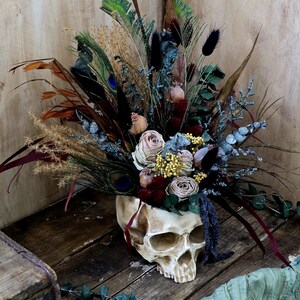 May include: A white skull vase filled with dried flowers, including roses, feathers, and eucalyptus. The vase is sitting on a wooden surface with a green fabric in the foreground.