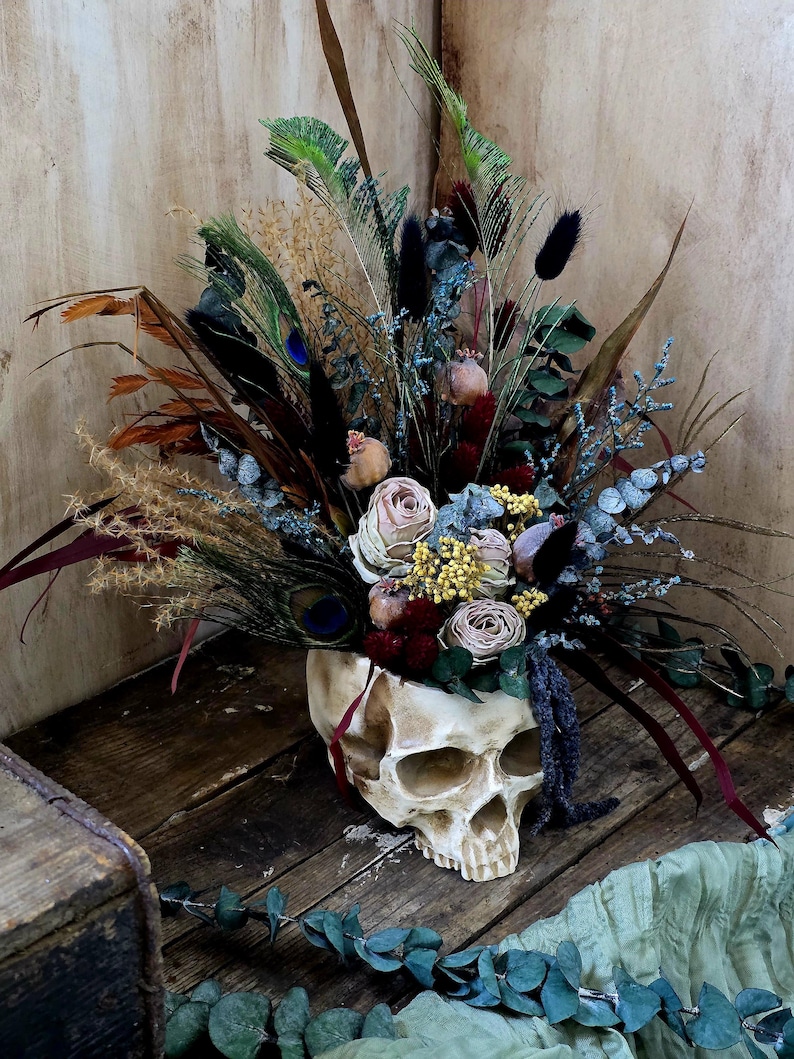 May include: A white skull vase filled with dried flowers, including roses, feathers, and eucalyptus. The vase is sitting on a wooden surface with a green fabric in the foreground.