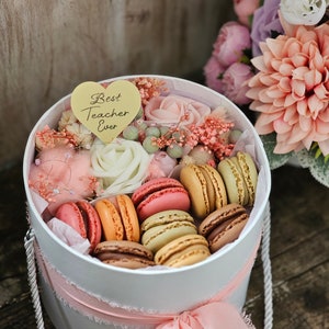 May include: A white gift box with a pink ribbon and a heart-shaped tag that says "Best Teacher Ever". The box is filled with macarons and dried flowers.