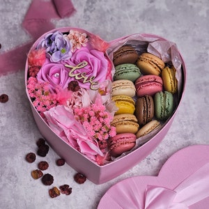 May include: A pink heart-shaped box filled with colorful macarons and pink, purple, and white flowers. The box has a matching lid with a pink satin bow. The word "Love" is written on a wooden tag.