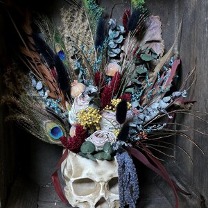 May include: A white skull planter filled with dried flowers and feathers. The flowers are in shades of pink, red, and blue. The feathers are black, green, and blue.