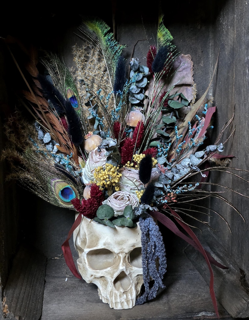 May include: A white skull planter filled with dried flowers and feathers. The flowers are in shades of pink, red, and blue. The feathers are black, green, and blue.