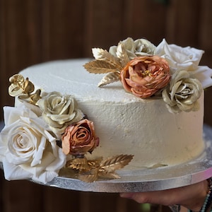 Op de afbeelding: Een ronde, geglazuurde taart versierd met witte, gouden en perzikroze bloemen en gouden bladeren. De taart staat op een zilveren taartbord. De taart is een dessert voor een bruiloft of een speciale gelegenheid.