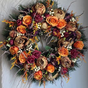 Könnte beinhalten: Ein kreisförmiger Kranz mit künstlichen Blumen in Orange, Burgunderrot und Braun. Der Kranz umfasst Rosen, Pfingstrosen und kleine weiße Beerenakzente, vor dem Hintergrund grüner Blätter. Ein dekoratives Stück für das Zuhause.