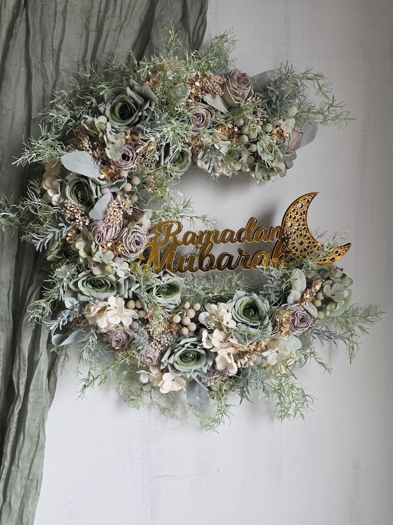 May include: A crescent-shaped floral wreath featuring green, white, and purple flowers, with gold-coloured leaves and a gold-coloured crescent moon. The words "Ramadan Mubarak" are written in gold.
