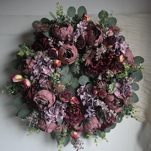 May include: A circular floral wreath with burgundy, mauve, and purple flowers, and green foliage. The wreath is densely packed with peonies, roses, and other blooms, creating a full and textured appearance. The wreath is a decorative item.