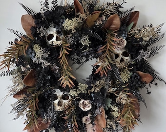 Può includere: Una ghirlanda circolare con tre teschi umani realistici incastonati tra fronde di felce nera, fogliame scuro, foglie di magnolia marroni e piccoli grappoli di bacche scure, arricchita da accenti floreali beige e rami sottili.