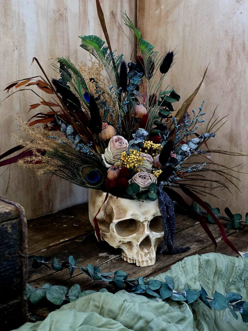 May include: A white skull planter filled with dried flowers, including roses, eucalyptus, and feathers. The skull is sitting on a wooden surface with a green fabric in the foreground.