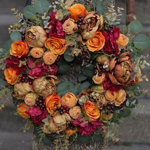 Op de afbeelding: Een herfstkrans met oranje, rode en bruine bloemen, groene bladeren en kleine donkere bessen.