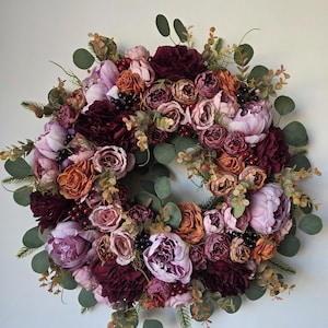 Può includere: Una ghirlanda floreale circolare caratterizzata da una densa disposizione di fiori artificiali nelle tonalità del bordeaux profondo, rosa antico, malva e arancione bruciato. Il design include foglie di eucalipto verdi, rametti di pino e bacche.
