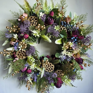 May include: A circular wreath composed of artificial flowers and foliage. It features deep purple roses, lavender, and green leaves, with gold-coloured berries and decorative pods. This wreath is suitable for seasonal or year-round decoration.