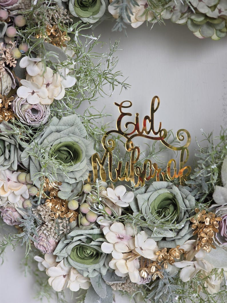 May include: A circular floral wreath with a gold 'Eid Mubarak' sign. The wreath is composed of green, white, and pink flowers, with gold and green accents. The flowers are arranged in a circular pattern, creating a decorative and festive item.
