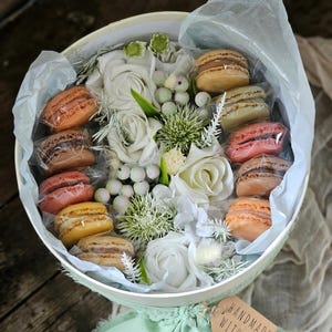 May include: A white box filled with white roses and macarons. The box is tied with a green ribbon and a brown tag that says "Handmade with love".