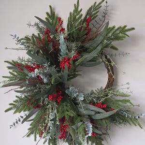 May include: A circular wreath constructed from a brown twig base, featuring a dense arrangement of green pine needles, long grey-green eucalyptus leaves, and clusters of bright red berries. The foliage is layered to create a full, textured appearance.