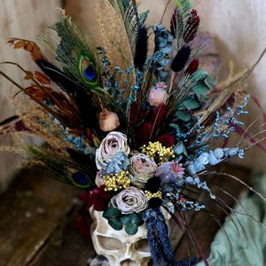 May include: A white skull vase filled with a bouquet of dried flowers, including roses, eucalyptus, and feathers. The flowers are in shades of blue, purple, and pink.