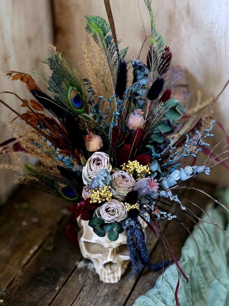 May include: A white skull vase filled with a bouquet of dried flowers, including roses, eucalyptus, and feathers. The flowers are in shades of blue, purple, and pink.