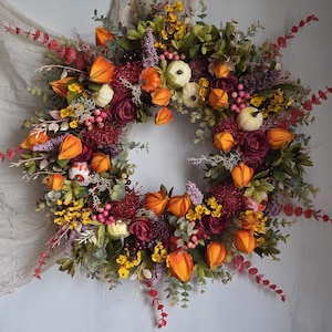 Pode incluir: Uma grinalda de outono circular com uma variedade de folhagens de outono. A grinalda apresenta lanternas chinesas laranja, pequenas abóboras brancas, rosas bordô e flores amarelas. Folhas verdes e bagas vermelhas completam o design.